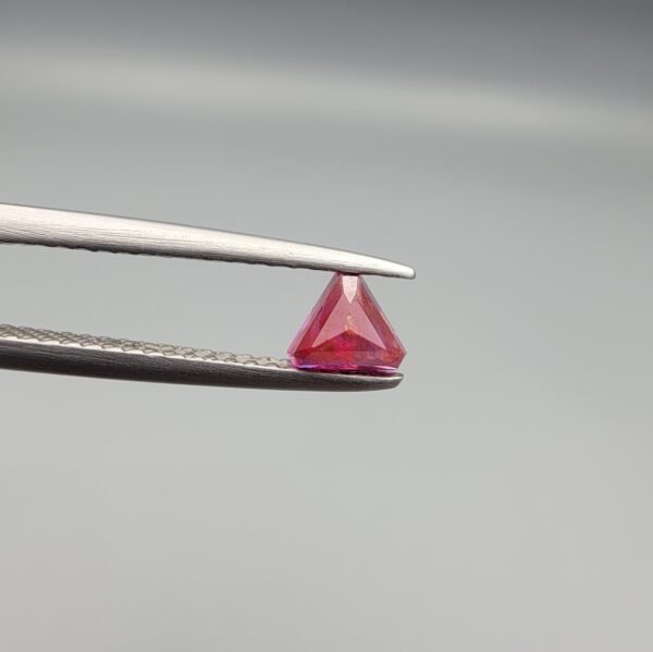 ​A 0.55 carat natural red Ruby (Yaqoot) from Kabul, Afghanistan, featuring a sharp triangle cut with rich color and natural internal silk, held in jeweler's tweezers.