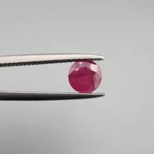 ​A 0.59 carat natural red Ruby (Yaqoot) from Kabul, Afghanistan, with a brilliant round cut, showcasing natural internal silk and vibrant saturation, held in jeweler's tweezers.