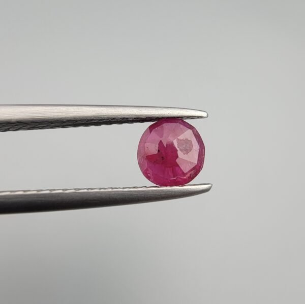 ​A 0.59 carat natural red Ruby (Yaqoot) from Kabul, Afghanistan, with a brilliant round cut, showcasing natural internal silk and vibrant saturation, held in jeweler's tweezers.