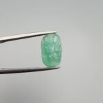Intricately hand-carved natural green emerald gemstone, 3.12 carats, featuring a floral motif from Zambia, held in professional jewelry tweezers.