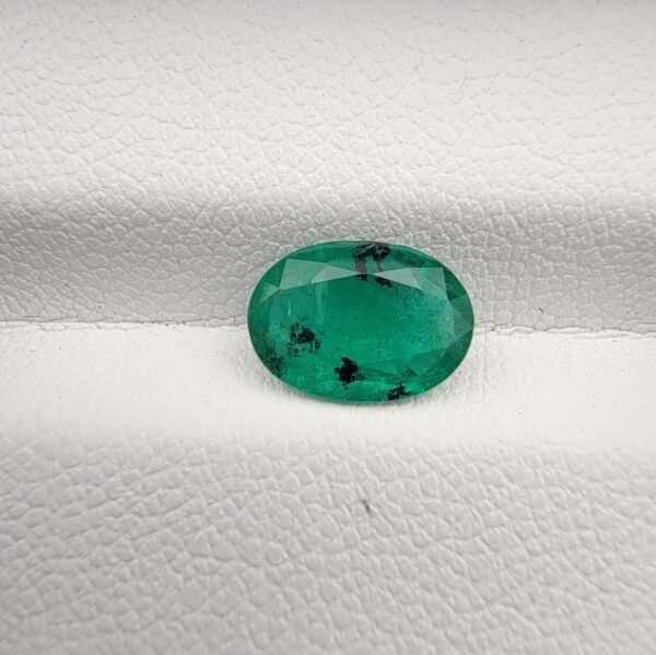Oval cut natural green emerald gemstone, 1.46 carats, sourced from Zambia, displayed on a white textured background.