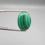 High-polished 21 carat natural malachite (Dana-e-Farhang) gemstone featuring deep emerald green vertical banding and a glossy oval cabochon finish, held in professional gemstone tweezers.