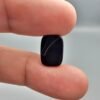 A 4.97 carat natural Black Agate (Sulemani Aqeeq) gemstone with a glossy oval cabochon cut, held in jeweler's tweezers against a dark background.