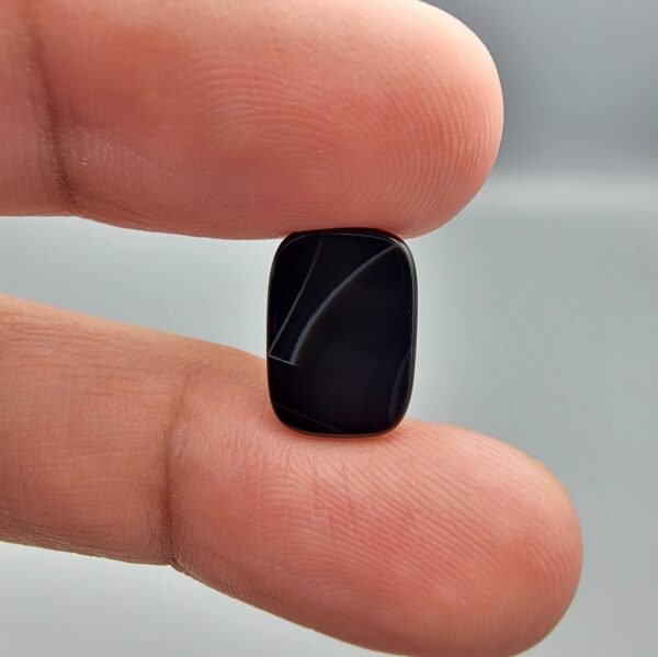 A 4.97 carat natural Black Agate (Sulemani Aqeeq) gemstone with a glossy oval cabochon cut, held in jeweler's tweezers against a dark background.