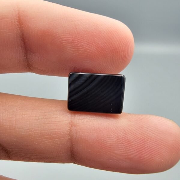 A 5.25 carat natural Black Agate (Sulemani Aqeeq) gemstone with an expertly polished oval cabochon cut, held in tweezers to showcase its high-luster surface and deep black color.