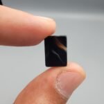 A 5.29 carat natural Black Agate (Sulemani Aqeeq) gemstone with a high-gloss oval cabochon cut, held in tweezers to showcase its dense black color and light-reflective surface.