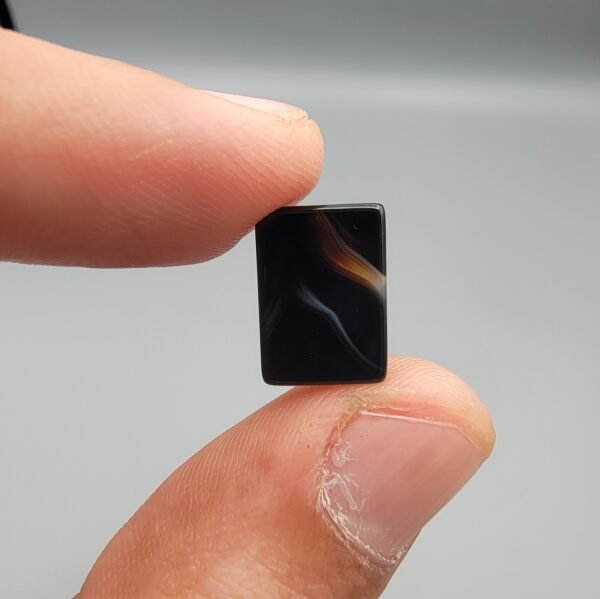 A 5.29 carat natural Black Agate (Sulemani Aqeeq) gemstone with a high-gloss oval cabochon cut, held in tweezers to showcase its dense black color and light-reflective surface.