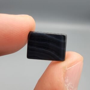 A 5.32 carat natural Black Agate (Sulemani Aqeeq) gemstone featuring a deep black, high-gloss oval cabochon cut, held in jeweler's tweezers against a dark professional background.