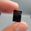 A 5.23 carat natural Black Agate (Sulemani Aqeeq) gemstone with a high-luster oval cabochon cut, held in tweezers to show its smooth, light-reflective surface and dense black color.