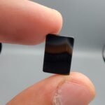 A 5.23 carat natural Black Agate (Sulemani Aqeeq) gemstone with a high-luster oval cabochon cut, held in tweezers to show its smooth, light-reflective surface and dense black color.