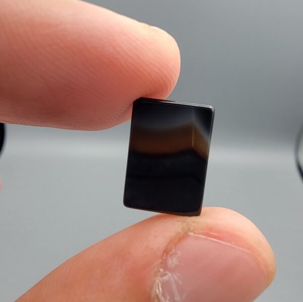 A 5.23 carat natural Black Agate (Sulemani Aqeeq) gemstone with a high-luster oval cabochon cut, held in tweezers to show its smooth, light-reflective surface and dense black color.
