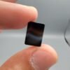 A 5.23 carat natural Black Agate (Sulemani Aqeeq) gemstone with a high-luster oval cabochon cut, held in tweezers to show its smooth, light-reflective surface and dense black color.