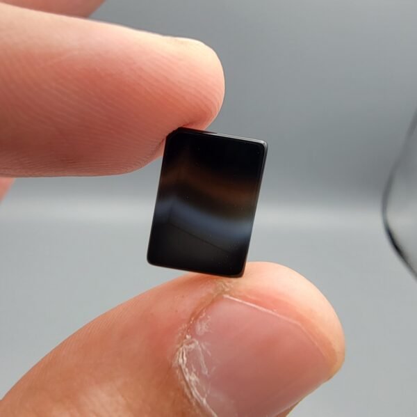 A 5.23 carat natural Black Agate (Sulemani Aqeeq) gemstone with a high-luster oval cabochon cut, held in tweezers to show its smooth, light-reflective surface and dense black color.