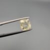 A 3.00 carat natural yellow sapphire (Pukhraj) held in tweezers, showing an oval cushion cut with visible mountain-like black inclusions on the reverse side against a grey background.