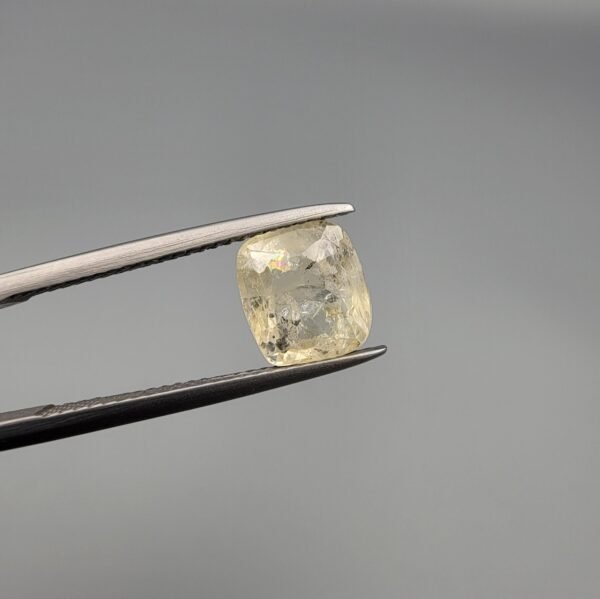 A 3.00 carat natural yellow sapphire (Pukhraj) held in tweezers, showing an oval cushion cut with visible mountain-like black inclusions on the reverse side against a grey background.