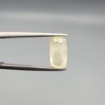 A 2.61 carat natural yellow sapphire (Pukhraj) held in tweezers, featuring a rare elongated cushion cut and a soft lemon-yellow hue against a neutral grey background.