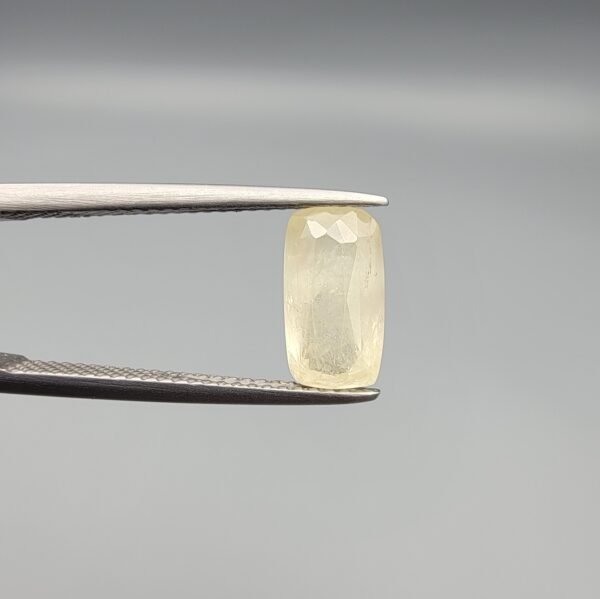 A 2.61 carat natural yellow sapphire (Pukhraj) held in tweezers, featuring a rare elongated cushion cut and a soft lemon-yellow hue against a neutral grey background.