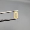 ​A 1.90 carat natural yellow sapphire (Pukhraj) held in jeweler's tweezers, featuring a rare elongated cushion cut and a soft lemon-yellow hue against a neutral grey background.