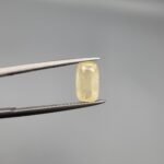 ​A 1.90 carat natural yellow sapphire (Pukhraj) held in jeweler's tweezers, featuring a rare elongated cushion cut and a soft lemon-yellow hue against a neutral grey background.