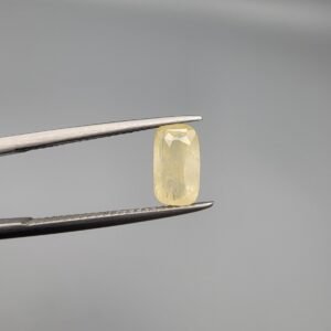 ​A 1.90 carat natural yellow sapphire (Pukhraj) held in jeweler's tweezers, featuring a rare elongated cushion cut and a soft lemon-yellow hue against a neutral grey background.