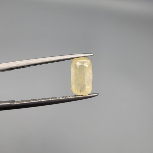 ​A 1.90 carat natural yellow sapphire (Pukhraj) held in jeweler's tweezers, featuring a rare elongated cushion cut and a soft lemon-yellow hue against a neutral grey background.