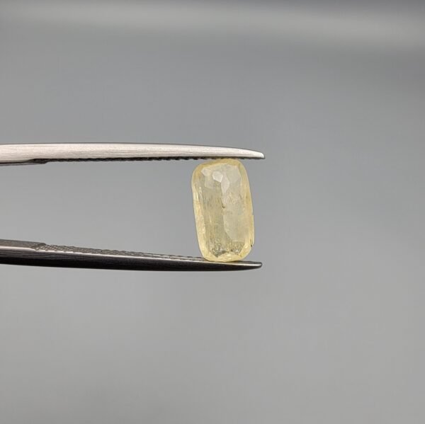 ​A 1.90 carat natural yellow sapphire (Pukhraj) held in jeweler's tweezers, featuring a rare elongated cushion cut and a soft lemon-yellow hue against a neutral grey background.