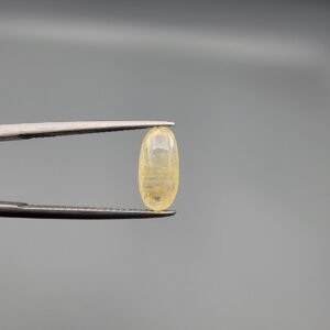 A 3.18 carat natural yellow sapphire (Pukhraj) held in jeweler's tweezers, featuring a rare elongated oval cut and a soft, transparent lemon-yellow hue against a neutral grey background.