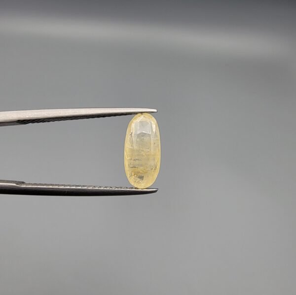 A 3.18 carat natural yellow sapphire (Pukhraj) held in jeweler's tweezers, featuring a rare elongated oval cut and a soft, transparent lemon-yellow hue against a neutral grey background.