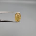 A 2.68 carat natural lemon yellow sapphire (Pukhraj) from Sri Lanka held in tweezers, featuring a brilliant oval cut and transparent clarity against a neutral grey background.