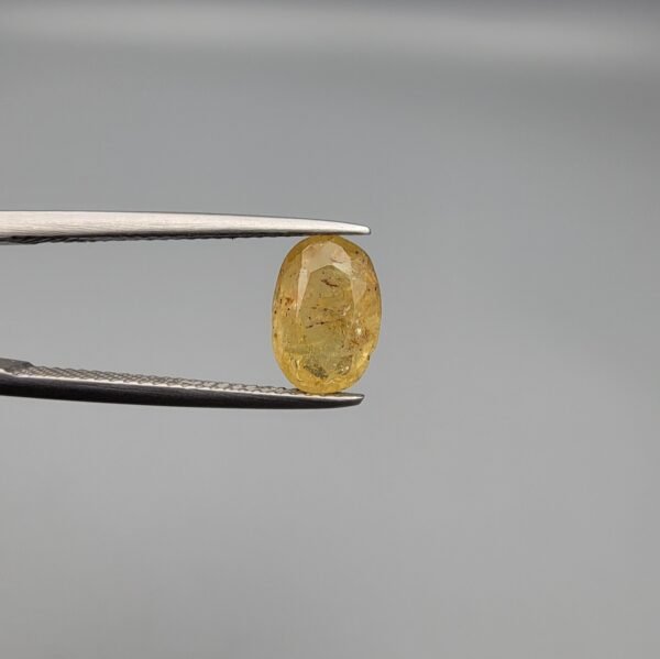 A 2.68 carat natural lemon yellow sapphire (Pukhraj) from Sri Lanka held in tweezers, featuring a brilliant oval cut and transparent clarity against a neutral grey background.