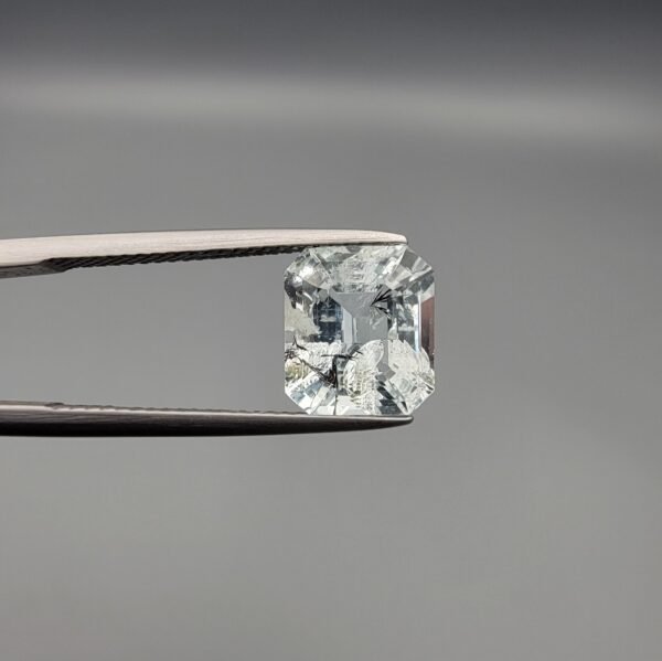 ​A 3.77 carat natural emerald-cut Aquamarine (Beruj) from Shigar Valley, Pakistan, displaying distinct black needle-like and "mountain" inclusions on the reverse side, held in jeweler's tweezers.