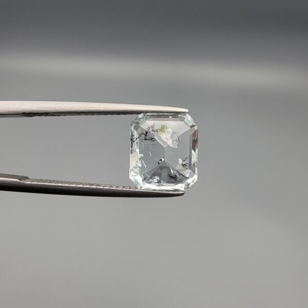 ​A 3.77 carat natural emerald-cut Aquamarine (Beruj) from Shigar Valley, Pakistan, displaying distinct black needle-like and "mountain" inclusions on the reverse side, held in jeweler's tweezers.