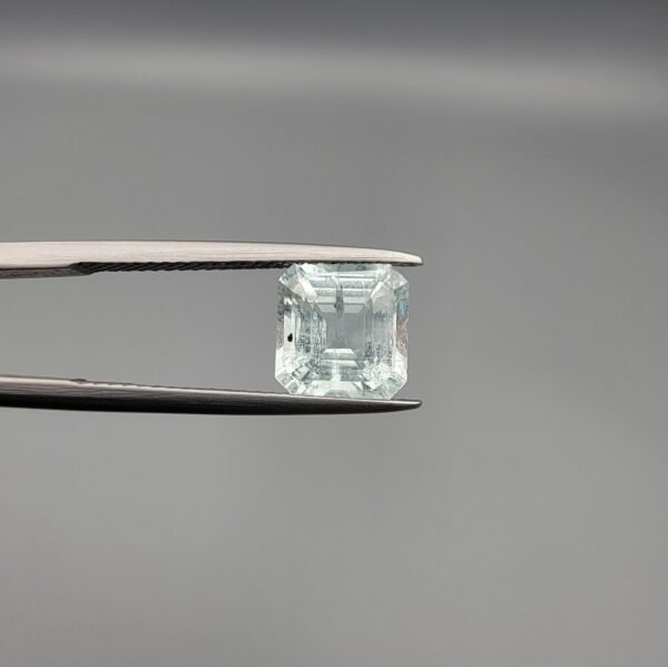 A 2.60 carat natural emerald-cut Aquamarine (Beruj) from Shigar Valley, Pakistan, held in tweezers, showing a clear face and a back side with distinct black needle-like mountain inclusions.