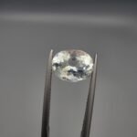 ​A 2.60 carat natural oval-cut Aquamarine (Beruj) from Shigar Valley, Pakistan, held in tweezers, displaying a clear face with distinct yellow mountain-like inclusions visible on the reverse.
