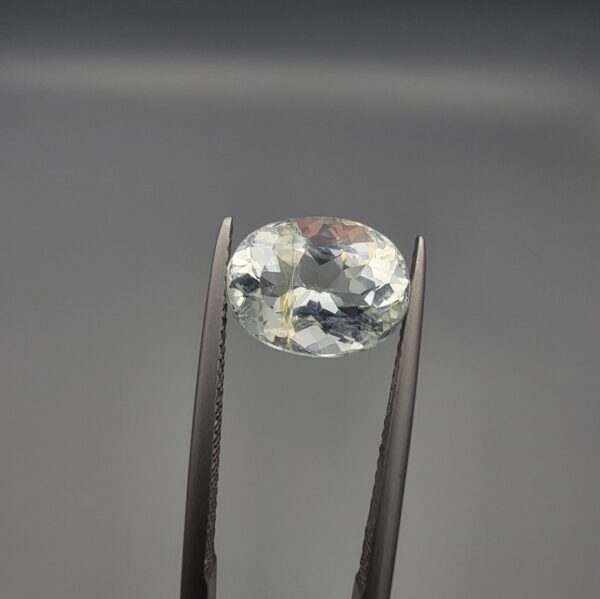 ​A 2.60 carat natural oval-cut Aquamarine (Beruj) from Shigar Valley, Pakistan, held in tweezers, displaying a clear face with distinct yellow mountain-like inclusions visible on the reverse.