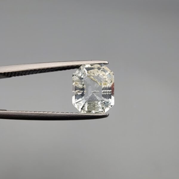 A 1.93 carat natural emerald-cut Aquamarine (Beruj) from Shigar Valley, Pakistan, held in tweezers, showing a clean face-up appearance with unique yellow and black mountain-like inclusions and natural surface pits on the reverse.