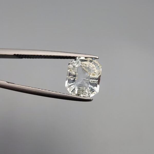 A 1.93 carat natural emerald-cut Aquamarine (Beruj) from Shigar Valley, Pakistan, held in tweezers, showing a clean face-up appearance with unique yellow and black mountain-like inclusions and natural surface pits on the reverse.