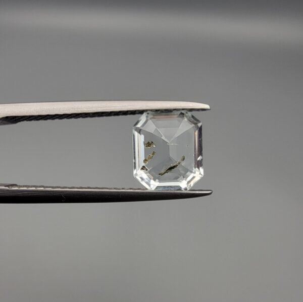 A 1.93 carat natural emerald-cut Aquamarine (Beruj) from Shigar Valley, Pakistan, held in tweezers, showing a clean face-up appearance with unique yellow and black mountain-like inclusions and natural surface pits on the reverse.