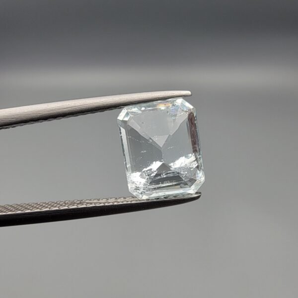 ​A 2.39 carat natural emerald-cut Aquamarine (Beruj) from Shigar Valley, Pakistan, held in jeweler's tweezers, showing a pale blue transparency with visible internal natural inclusions.