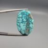 A 25-carat natural Iranian Turquoise (Feroza) from Kerman City, featuring a large oval cabochon cut with a vibrant blue-green base and dense golden pyrite matrix, held in jeweler's tweezers.