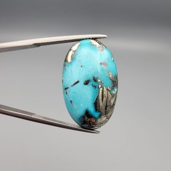 A 23-carat natural Iranian Turquoise (Feroza) gemstone with a smooth oval cabochon cut, displaying a vibrant sky-blue color and metallic pyrite inclusions, held in tweezers.