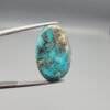 A 23-carat natural Iranian Turquoise (Feroza) gemstone with a smooth oval cabochon cut, displaying a vibrant sky-blue color and metallic pyrite inclusions, held in tweezers.