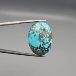 A 37.23-carat natural Iranian Turquoise (Feroza) from Kerman City, featuring a substantial oval cabochon cut with a vibrant sky-blue base and a heavy golden pyrite matrix, held in jeweler's tweezers.