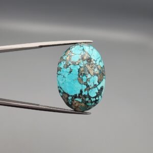 A 37.23-carat natural Iranian Turquoise (Feroza) from Kerman City, featuring a substantial oval cabochon cut with a vibrant sky-blue base and a heavy golden pyrite matrix, held in jeweler's tweezers.