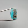 ​A 20.58-carat natural Iranian Turquoise (Feroza) from Kerman City, featuring an oval cabochon cut with a clear blue base and natural "golden" pyrite inclusions, held in jeweler's tweezers.