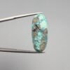 A 14.58-carat natural Iranian Turquoise (Feroza) from Kerman City, featuring an elegant oval cabochon cut with a vivid blue body and subtle metallic pyrite flecks, held in jeweler's tweezers.