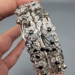 Pair of handcrafted pure silver bangles featuring a floral vine design set with black and white zircons, weighing 52.31 grams, held against a grey background.
