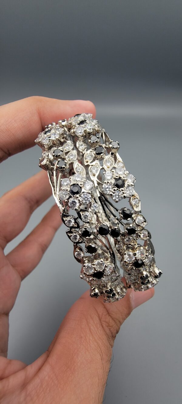 Pair of handcrafted pure silver bangles featuring a floral vine design set with black and white zircons, weighing 52.31 grams, held against a grey background.