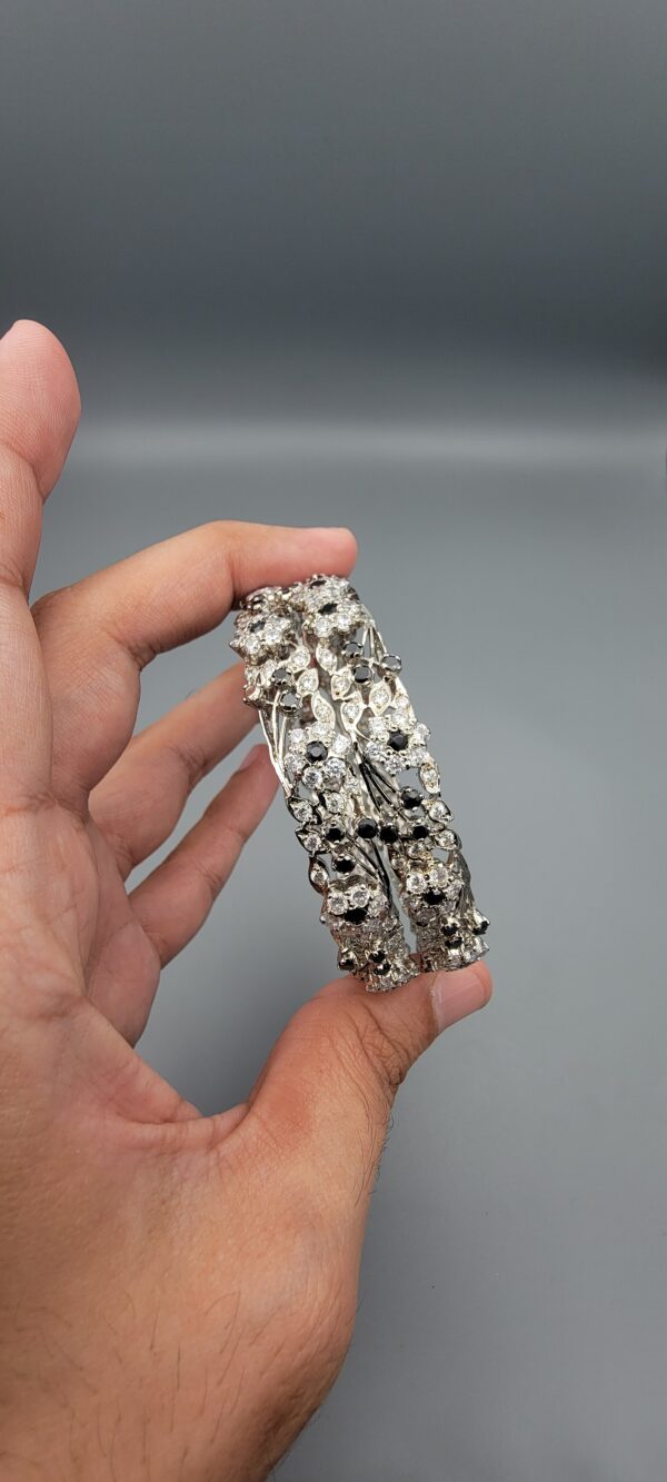 Pair of handcrafted pure silver bangles featuring a floral vine design set with black and white zircons, weighing 52.31 grams, held against a grey background.