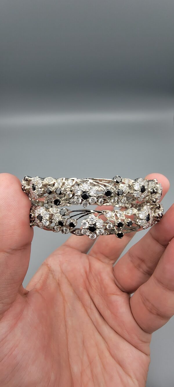 Pair of handcrafted pure silver bangles featuring a floral vine design set with black and white zircons, weighing 52.31 grams, held against a grey background.